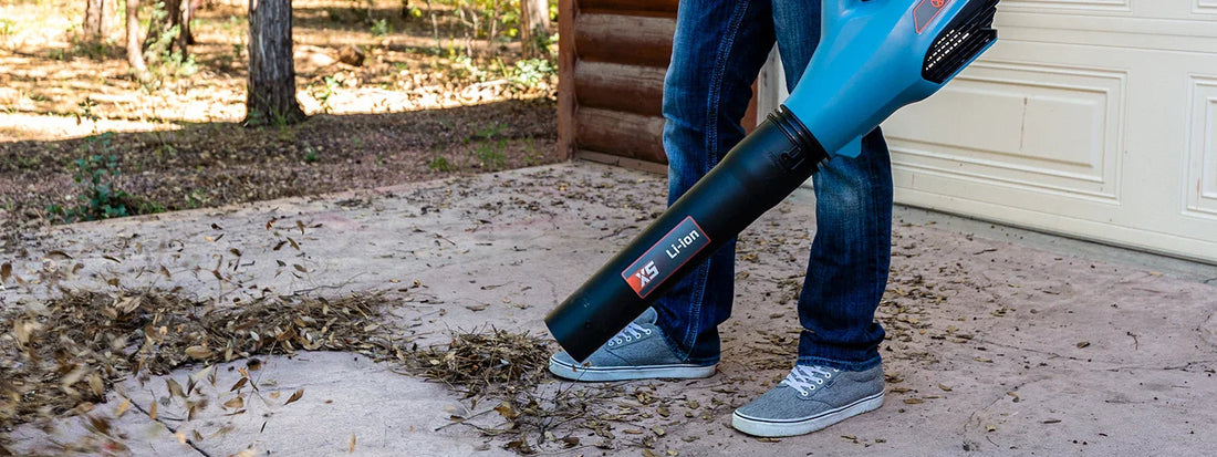 Person using blue electric leaf blower with XS Li-ion label to clear dry leaves on concrete driveway near wooden house