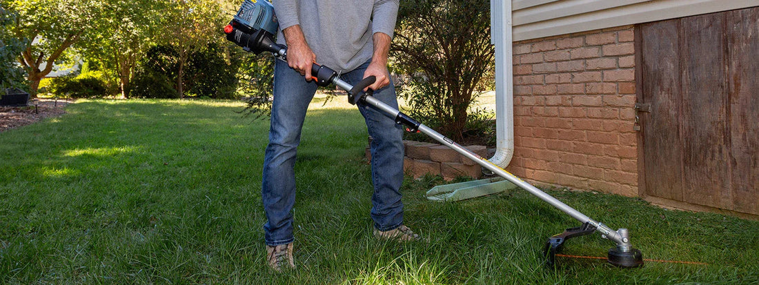 Person using a gas string trimmer to cut grass next to a brick house in a sunny backyard