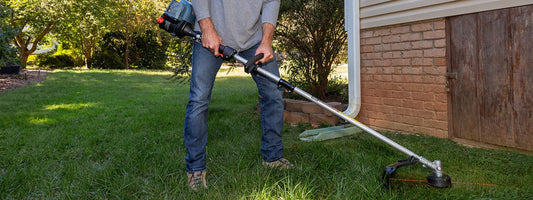 Person using a gas string trimmer to cut grass next to a brick house in a sunny backyard