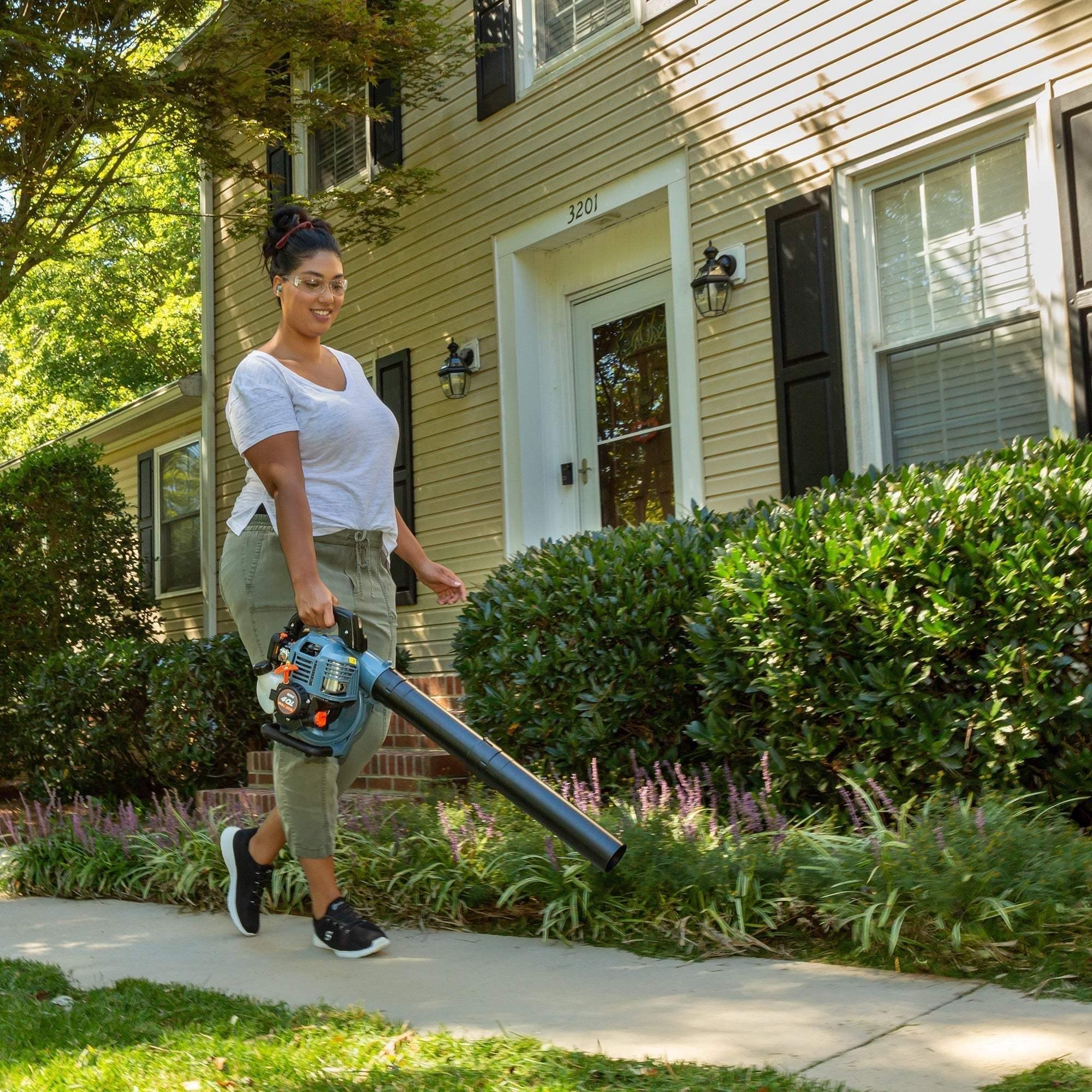 Woman using a handheld leaf blower to clear sidewalk near house with bushes
